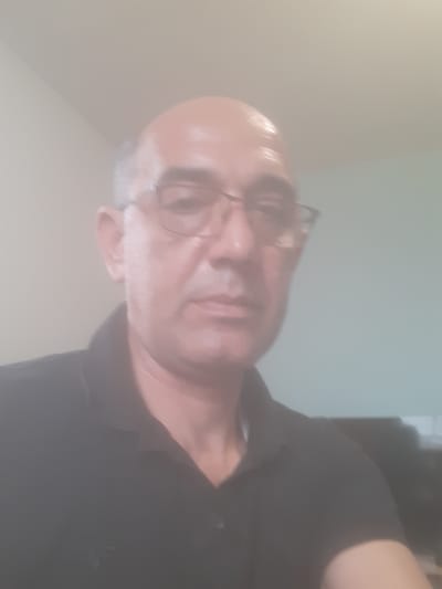 The image shows a bald man wearing glasses and a black shirt. The background is blurry and suggests an indoor setting with light-colored walls and possibly a television or monitor. There is no information to determine the location is Dolni Chiflik, Bulgaria.
