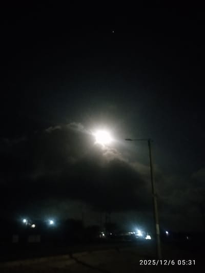 A nighttime landscape features a bright moon obscured by dark clouds, casting a diffused glow. A streetlight stands on the right, its beam contrasting with the moonlight. Below, a road leads into the distance where faint lights suggest buildings or vehicles. The darkness dominates, punctuated by the bright moon and scattered lights. The date "2025/12/6 05:31" is printed at the bottom.
