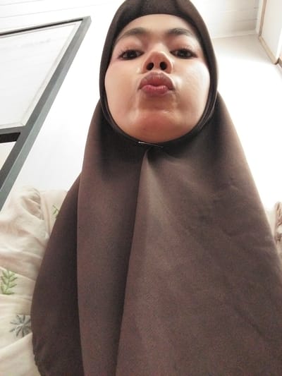 This is a selfie taken by a young woman wearing a brown hijab. She is posing with her lips puckered, looking directly at the camera. Her face is close to the lens, with emphasis on her eyes and lips. She has well-defined eyebrows and her eyelashes appear to be enhanced with mascara. Her skin is smooth and appears to have a light foundation applied.

The background of the image is partially visible and appears to be an indoor setting. To the left, a white wall with horizontal paneling can be seen, along with the top of a wooden structure. In the lower part of the image, a portion of a picture frame with a gray border is visible, suggesting the woman might be near a mirror or a framed photograph. There is also a glimpse of white fabric with subtle green embroidered patterns beneath the frame, possibly a piece of clothing or bedding.

The lighting suggests an indoor environment, possibly during the daytime given the brightness. There are no obvious external cues to indicate the time of day or weather conditions. The woman's expression is playful and flirtatious, indicated by her pouting lips and direct gaze. No visible text is present in the image. The context provided places the location as Tibojong, Indonesia.