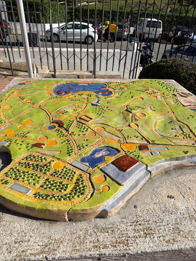A high-angle, slightly tilted shot presents a park map in Arroyo De La Miel-Benalmadena Costa, Spain, mounted on a wall behind a black metal fence. The map, made of textured materials like paint and possibly tile, depicts a green park landscape with winding yellow paths, representations of bodies of water (blue), tree clusters (green dots), buildings (brown and grey rectangles), and circular areas. The surface on which the map is mounted is textured concrete. Outside the fence, a view of a street includes parked cars, a small green bush, and parts of buildings. The primary subject is the park map itself. The day appears sunny, casting shadows and highlighting the map's texture.