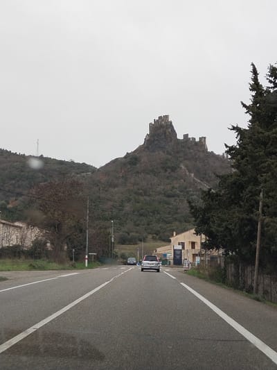 The image depicts a road scene in Rochemaure, France, under an overcast, light grey sky. A paved, two-lane road with solid white fog lines and a dashed white center line occupies the foreground and recedes into the distance. Visible wet patches are present on the asphalt. A silver or light-colored SUV is centrally positioned on the road, facing away from the camera, followed by a darker vehicle further down the road.

To the right of the road, a dense, dark green coniferous tree partially obscures a two-story, light-colored building with multiple signs affixed to its exterior. Further right, a fence line runs parallel to the road. On the left side, a light-colored, stone-faced building is partially visible behind sparse deciduous vegetation. Utility poles and overhead power lines are visible along the road and extending up the hillside on the left.

The background is dominated by a rugged, rocky mountain largely covered with brown and green vegetation, indicating a cool season. Atop this mountain, the stone ruins of the Château de Rochemaure are prominently situated, displaying irregular walls, towers, and battlements. Another utility tower is visible on a lower hill to the far left. No individuals are visible in the scene.