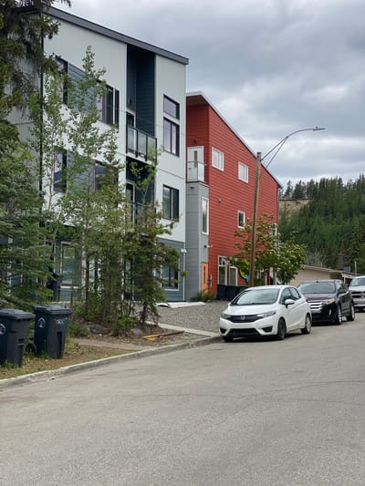 A residential street in Whitehorse, Canada, is depicted under an overcast sky. The image features a multi-story, modern building with a gray and teal facade on the left, and a striking red addition on the right. Trees are sparsely planted in front of the building, and two large black trash bins are visible on the left side of the image. A white Honda Fit is parked in front of the building, followed by a black SUV and a silver pickup truck. The street is paved, with a concrete curb and a gravel area bordering the building. In the background, a treed hillside rises, with some erosion visible at its crest. A street lamp arches over the scene.