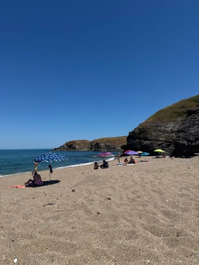 This is a wide shot of a sandy beach in Sinemorets, Bulgaria, under a clear blue sky. The foreground is dominated by the sandy shore, with scattered shells and debris. In the mid-ground, several beach umbrellas are set up, offering shade to people sunbathing or relaxing on towels. Some individuals are seated, while others appear to be lying down. A few people are visible near the water's edge, with one person in the sea. The water is a calm, blue-green. In the background, rocky cliffs covered in dry grass rise from the shoreline, forming a natural backdrop to the beach. The overall scene depicts a serene day at the beach.