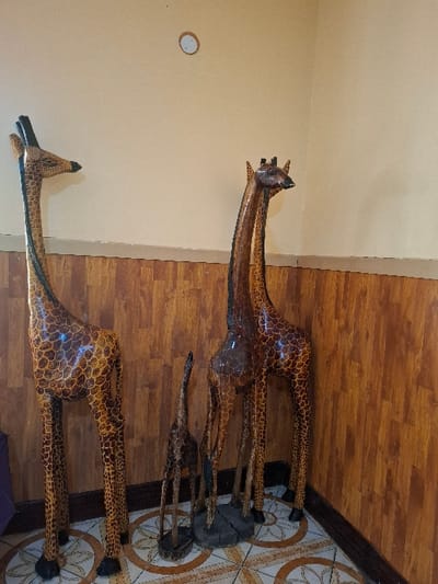 The image shows three wooden giraffe sculptures in a room with brown wood-panelled walls. The sculptures stand on a tiled floor. The sculptures are all different sizes, with the largest one standing on the left side of the image and the smallest one standing on the right. The two larger sculptures have long necks and heads with long, pointed ears. The smallest sculpture has a shorter neck and head and lacks any discernible detail. All three sculptures are carved in detail to resemble giraffes, with a brown and black patterned paint job. The sculptures are in a room located at the coordinates -0.754, 36.438, which is in Karagita, Kenya.