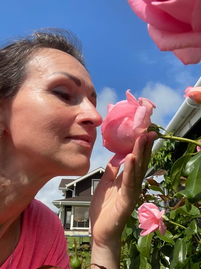 A woman is shown from the side, smelling a pink rose in a garden. She is wearing a pink t-shirt and has dark hair. The background includes a house and a bright blue sky with some clouds. The woman's face is illuminated by sunlight, and there are visible skin textures and fine lines. A hand is holding the rose stem, and other pink roses and green leaves are visible on the plant. The location is identified as Solov'evka, Russia.