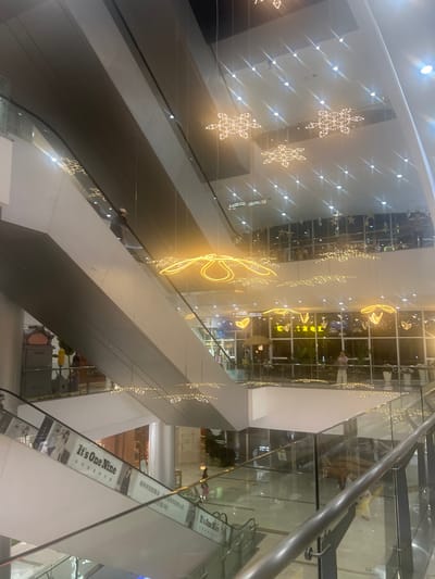 The image depicts the interior of a multi-level shopping mall, likely in Liandu District, China, characterized by escalators, glass railings, and ornate lighting fixtures. Several escalators are visible, some ascending and others descending, with advertisements displayed along their sides. People are seen riding the escalators and walking on the different levels. The mall's ceiling is adorned with numerous snowflake-shaped and flower-shaped light installations, creating a festive and illuminated atmosphere. Large windows on the upper levels offer glimpses of a cityscape with more lights, suggesting it is evening or night. The architecture appears modern, with clean lines and reflective surfaces.