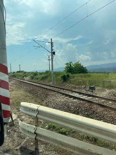 This image presents a daylight scene of an electrified railway line in a rural setting, located in Karnare, Bulgaria. The vantage point appears to be low, possibly from a vehicle or adjacent to a road.
In the immediate foreground, to the left, a portion of a red and white striped pole is visible, consistent with a railway crossing barrier or marker. Across the lower part of the frame, a two-tiered, weathered, off-white metal guardrail is present. The ground in this area consists of dry, sparse grass and bare soil.
The midground features two parallel railway tracks, laid on a bed of crushed stone ballast, extending diagonally from the lower-left towards the middle-right. A faint wisp of white smoke or steam is visible rising from the track area in the central-left midground. To the right of the tracks, a series of green bushes and trees line the railway corridor. A tall concrete utility pole equipped with a catenary support structure and insulators stands to the right of the tracks, bearing a white sign with the numbers "132" and "7". Further to the right and slightly behind, a chain-link fence separates the railway area from a grassy field. A dark, rectangular metal box on a stand is positioned near the tracks on the right.
The background shows expansive green fields leading to distant, hazy mountains or hills on the horizon. The sky is partly cloudy, displaying light blue areas with scattered white cumulus clouds. Overhead electrical lines, part of the railway's catenary system, are visible against the sky. No individuals or active actions are depicted, and no trains are visible on the tracks.