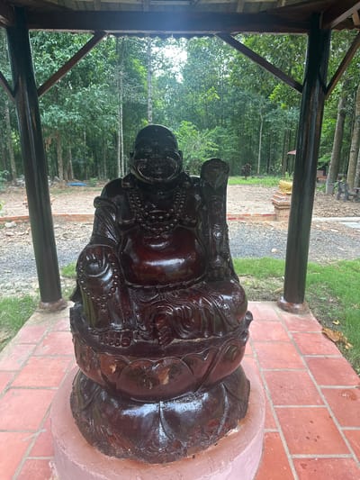 This media file captures a large, dark brown, carved wooden statue of a laughing Buddha, likely intended as a decorative garden ornament. The statue is positioned outdoors on a raised, pinkish-colored pedestal, which is surrounded by red brick paving. The Buddha statue is elaborately carved, featuring intricate details in its facial expression, clothing, and ornamentation, including a beaded necklace.
The setting appears to be a tranquil, possibly religious or spiritual garden space. Behind the statue, a dense forest of green trees and vegetation is visible, suggesting a rural or semi-rural location. The ground in the background is a mix of grass, gravel, and dirt, with some scattered bricks. A wooden structure, possibly part of a gazebo or shelter, is visible in the foreground, with dark wooden posts and beams providing a framing element.
The lighting suggests it is daytime, with soft natural light illuminating the scene. There are no visible signs of specific activities or events taking place; the image appears to be a static shot of the statue within its environment. The overall mood conveyed is one of peace and serenity, befitting the subject matter of the Buddha. There is no visible text in the image. The location context provided is Suối Câu, Vietnam.