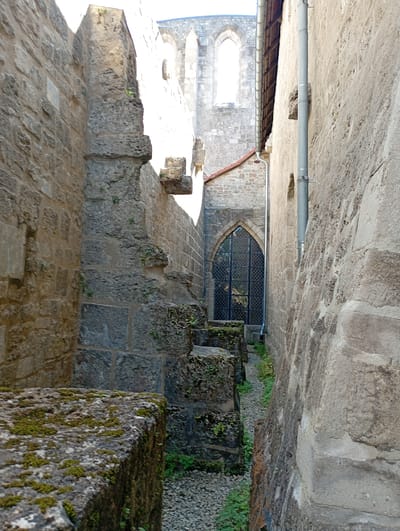 The image shows a narrow alleyway between two stone walls, leading towards a gothic arched doorway with a latticed window. The walls are made of rough-hewn stone blocks, some of which are covered in moss. In the foreground on the left, there is a large, moss-covered stone block. On the right, the wall is a lighter, smoother stone. In the background, part of a larger stone building with arched windows is visible, suggesting it could be a church or monastery. The alleyway is covered with gravel and some sparse green vegetation. The scene appears to be part of the ruins of the Walkenried Abbey in Walkenried, Germany, known for its Romanesque and Gothic architecture. There are no people or animals visible in the image. The lighting suggests it is daytime, with sunlight illuminating parts of the scene.