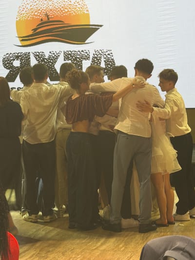 This media file depicts a group of young people, appearing to be in their late teens or early twenties, engaged in a collaborative dance or performance. The subjects are dressed in casual attire, with several individuals wearing white shirts and light-colored pants or skirts, suggesting a cohesive costume theme. One person is wearing a dark velvet top.

The scene appears to be indoors, possibly on a stage or in a studio, with a large banner or backdrop behind the performers. The banner features a stylized graphic of a sun or sunset over water, with dark, abstract shapes and what looks like the word "Khotianivka" printed in large, bold, black letters. The lighting is warm and somewhat dim, casting shadows and creating a dramatic atmosphere. The overall mood seems focused and dynamic, with the performers intertwined in various poses, indicating movement and interaction.

The arrangement of the individuals suggests a choreographed routine, possibly a contemporary dance or a theatrical piece. Some individuals are supporting others, creating a visual sense of unity and effort. The setting hints at a public performance or a rehearsal space within Khotianivka, Ukraine, given the visible text on the backdrop. No specific time of day or weather conditions are discernible from the indoor setting. The focus is clearly on the human figures and their collective activity.