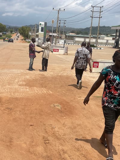 The image depicts a scene in Unguwar Saya, Nigeria. Two men are shaking hands in the foreground, with one wearing a purple and red striped shirt and blue jeans, and the other in a light-colored shirt and dark pants. Behind them, a man in a patterned shirt walks away from the camera, and a person in a floral-patterned top walks towards the camera. In the background, there is a gas station with a sign indicating "NNPC" and fuel prices. Power lines and utility poles crisscross the sky, with hills visible in the distance under a cloudy sky. The ground is unpaved and appears to be reddish-brown dirt. Several vehicles, including cars and motorcycles, are on the road in the middle ground, suggesting an active urban or semi-urban environment.