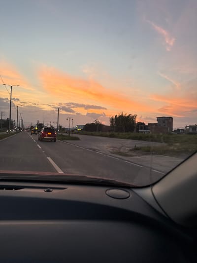 The image depicts an exterior scene from the perspective of a vehicle's interior, looking forward along a road at sunset in Pitalito, Colombia.

**Foreground:** The dark-toned dashboard and lower edge of a windshield are visible across the bottom of the frame, indicating the vantage point is from inside a car.

**Midground:** A two-lane paved road extends into the distance, marked with white dashed lane lines. A red SUV, possibly a Ford Explorer, is directly ahead in the left lane with its taillights illuminated. Further ahead, a smaller vehicle, likely a motorcycle or mototaxi, is visible in the right lane. Utility poles with overhead lines and streetlights line the left side of the road. On the right side, a grassy verge separates the road from an undeveloped area.

**Background:** The horizon reveals a mix of undeveloped land, sparse vegetation, and structures. A cluster of bamboo-like trees is present on the right. Beyond them, a multi-story, unfinished brick building with an open top level stands prominently. Other smaller structures and a commercial billboard displaying the text "Mall" are also discernible on the right. The sky dominates the upper portion of the scene, showcasing a sunset with broad, horizontal bands of orange and pink clouds transitioning into a lighter blue overhead.