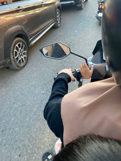 This media file shows a close-up perspective from someone riding on a vehicle, possibly a motorcycle, in Ta', Indonesia. The primary subjects are two individuals, an adult and a child, whose hands are visible. The adult's hands are on the handlebars of the vehicle, while the child's smaller hands are also near the handlebars, suggesting they are a passenger. The adult is wearing a black long-sleeved shirt, and the child is wearing a white long-sleeved shirt beneath a light brown garment.

In the background, a dark-colored car is visible, as is a portion of the road surface. A side mirror of the vehicle the subjects are on reflects the road and the car. The scene appears to be an urban or suburban environment due to the presence of vehicles and the paved road.

The image seems to have been taken during daylight hours, as indicated by the lighting conditions. There are no strong cues about the weather, but the lighting suggests it's not dark or raining heavily. The overall mood is difficult to ascertain from the cropped image, but the presence of a child suggests a family outing or travel.

No discernible text is visible in the image. The focus is on the interaction between the adult and child and the act of traveling together.