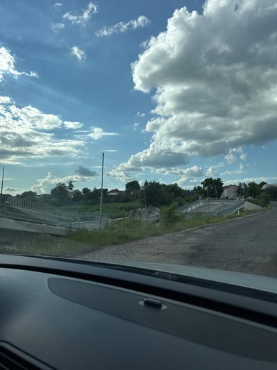 The media file displays a scene captured from within a vehicle, looking outwards. The primary subjects are a bright blue sky filled with a mix of large, dramatic cumulus clouds and smaller, wispy cirrus clouds. The sunlight filtering through the clouds creates a dynamic contrast, suggesting it is daytime, likely mid-morning or afternoon.
The scene outside the vehicle shows a slightly elevated view of a rural or suburban environment. A paved road runs along the right side of the frame, with a grassy verge and vegetation. There appears to be a metal railing or fence structure running parallel to the road and a body of water or a canal. Beyond this, trees and a few buildings, possibly houses, are visible, nestled amongst greenery. The overall impression is one of a clear, bright day, with the weather appearing fair.
There is no discernible activity or event taking place beyond the passive observation of the landscape. No people are visible. Notable details include the texture of the road, the lushness of the vegetation, and the varied cloud formations. The interior of the vehicle is partially visible on the right side of the frame, showing a dark grey surface with a textured speaker grille and some indistinct lines. No visible text is present in the media file. The location context is noted as unknown, Ukraine.