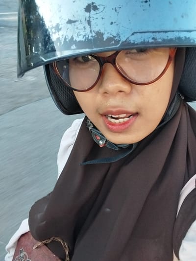 A woman wearing a blue helmet and glasses is captured in a close-up selfie while riding a motorcycle. She is also wearing a brown hijab and a white shirt. Her mouth is slightly open, revealing her teeth. The background suggests an outdoor setting with a blurred, gray surface that could be a road. The image is taken from a low angle, looking up at the woman.