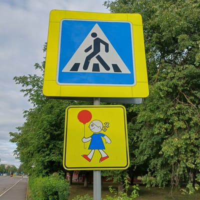 Two rectangular road signs with prominent yellow reflective borders are affixed vertically to a gray metal pole in Baranavichy, Belarus. The upper sign consists of a blue square panel with a white upward-pointing triangle, inside which a black silhouette of a pedestrian walks across a depicted crosswalk. The lower sign, also square with a yellow reflective border, features a yellow reflective background on which a stylized white-skinned child figure, wearing a blue dress and red shoes, with distinctive white hair styled in pigtails, holds a red balloon. To the left of the pole, a paved sidewalk parallels a road where distant vehicles and buildings are discernible under an overcast sky. The right and upper background are densely populated with vibrant green foliage from mature deciduous trees, with some visible branches.