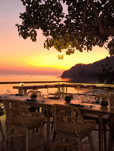 This media file depicts a tranquil outdoor dining setting overlooking a body of water at sunset. In the foreground, a wooden table is set with place settings, including glassware, napkins, and cutlery. Woven rattan chairs surround the table, suggesting a comfortable and inviting atmosphere.
The scene is set against a breathtaking sunset, with the sky transitioning through hues of pink, orange, and yellow. The calm water reflects the vibrant colors of the sky, creating a serene and picturesque view. A lush green tree with dark leaves hangs over the dining area, casting dappled shadows and adding a natural element to the composition. A decorative woven lamp hangs from a branch of the tree, illuminated by the fading light.
In the background, a hilly landscape with a pier extending into the water is visible. The overall mood is peaceful and romantic, ideal for a leisurely dinner or special occasion. The time of day is clearly sunset, given the colors of the sky and the low angle of the sun. The location appears to be a coastal area, possibly a restaurant or resort, given the organized dining setup and the scenic view. There is some visible text on a small black sign on the table, which appears to read "Ibusita".
