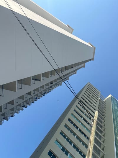 The image presents a low-angle view of two tall buildings against a clear blue sky, likely taken in daylight conditions.
The building on the left is white and features numerous balconies arranged vertically along its facade. The architectural style is simple and repetitive. The building on the right is taller and appears to be more modern. It is light beige, with a repetitive grid of windows. A wooden power pole obscures some of the windows. Wires stretch from the buildings and traverse the sky, intersecting the view.
A couple of birds can be seen in the sky between the buildings.
Based on the composition and architectural styles, it is likely the image was taken in Acapulco, Mexico.