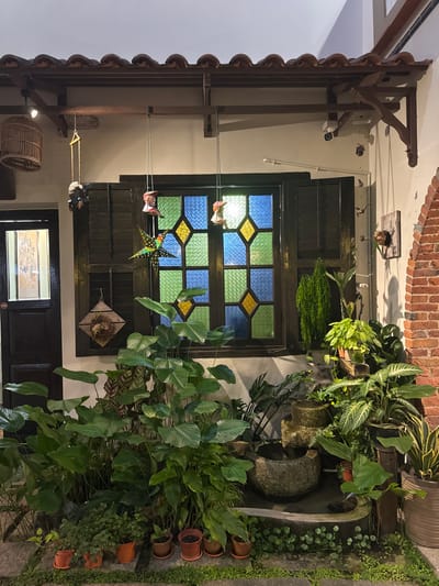 The image displays an exterior or semi-outdoor architectural facade in George Town, Malaysia, characterized by a blend of traditional and decorative elements with extensive flora. A terracotta-tiled roof overhang is supported by dark wooden beams and brackets. Below this structure, a white wall features a large, dark-framed window filled with stained glass panes arranged in a pattern of blue and green rectangles accented with yellow diamonds. To the left of the window, a dark wooden door with a glass panel is visible. On the far right, a section of exposed red brick wall forms an arch-like structure.
Decorative objects are suspended from the roof overhang and affixed to the walls. A wooden birdcage hangs on the far left. Adjacent to it, a small, dark-colored figure, resembling a monkey, swings from a rope. In front of the stained glass window, two colorful, bell-shaped ornaments and a larger, multi-colored star-shaped ornament are suspended. A square wooden plaque adorned with a woven wreath is mounted on the wall to the left of the window. A white security camera and a smaller dark sensor are affixed to the white wall beneath the roof overhang, above the window. On the right brick wall, a small wooden carving of a bird or similar figure is mounted.
The lower portion of the scene is dominated by dense greenery. Large-leafed green plants form a prominent cluster beneath the window. In the foreground, several smaller potted plants in terracotta pots are arranged on the ground, which is partially covered by trailing green vegetation and paving stones. To the right, a multi-tiered stone water feature or fountain is nestled among various potted plants, including tall, slender species, trailing plants, and those with variegated leaves. The entire scene is illuminated by artificial light. No individuals are present.
