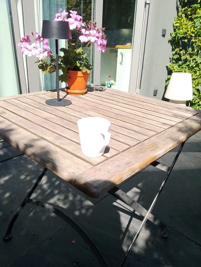 The image depicts an outdoor scene, likely a patio or garden, with a wooden table in the foreground. The table has a weathered appearance, with visible wood grain and plank construction. On the table, there is a white ceramic mug positioned towards the right side. To the left, a black lamp with a conical shade stands next to a potted plant with pink flowers. The plant is in an orange terracotta pot. In the background, a building with large windows and ivy-covered walls is visible. A second, white lamp with a similar conical shade is situated further to the right. The ground beneath the table appears to be dark stone or tile, with shadows from the table legs and surrounding foliage cast upon it. A few pink flower petals are scattered on the ground. The scene suggests a relaxed, possibly sunny day. The location is specified as Winkel, Germany.