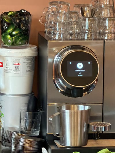 This close-up image depicts a beverage and dessert preparation station in Villecresnes, France. A stainless steel commercial Nespresso coffee machine is prominently positioned on the right, featuring a central circular digital display that reads "insérer la capsule" (insert the capsule) and "eau chaude" (hot water) alongside a Nespresso logo and water droplet icon. A stainless steel milk frothing pitcher sits on the machine's black drip tray beneath the dispenser, with a chrome steaming wand visible to its right. Multiple clear glass espresso-style cups are stacked on top of the Nespresso machine. To the left, a clear plastic container filled with green and dark-colored Nespresso coffee capsules is stacked on top of a white 3kg Nutella bucket, labeled with Ferrero branding and French text for professional recipes. Below the Nutella bucket, a silver cylindrical container with partial "MÖVENPICK" branding is visible. On the dark countertop in the bottom left, a stack of multiple silver-colored spoons is present next to a clear glass cup, also faintly branded "MÖVENPICK," which holds a dark utensil. The background consists of a plain, light brown wall.