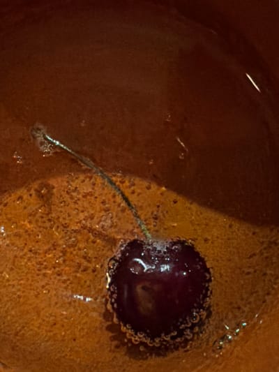 A single cherry with a stem is submerged in a reddish-brown liquid within a bowl. The liquid has a granular texture and contains small bubbles, particularly around the cherry. The background is a smooth, dark reddish-brown surface, possibly the interior of the bowl. There are no people or indications of a specific location in Shabdan, Kyrgyzstan.