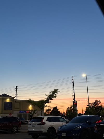 The image depicts a dusk scene in a commercial area of Vaughan, Canada. The sky transitions from a clear blue at the top to a vibrant orange and pink hue near the horizon, indicating sunset. Power lines crisscross the frame, supported by utility poles. A street lamp is illuminated, casting a bright light.
In the foreground, several vehicles are parked, including a dark-colored SUV on the left, a white SUV in the center, and a dark blue electric vehicle on the right. The license plate of the blue electric vehicle is visible, reading "CVEF-780".
A building with illuminated signage is visible on the left. One sign reads "MERRIAM SCHOOL OF MUSIC". Another sign above a storefront appears to say "MERRIAM piano". A red "STOP" sign is also visible. To the right of this building, a tall pylon with multiple signs is lit up, featuring names such as "MEGABITE" and "DIAMOND NAILS".
Trees, some with foliage, are scattered throughout the scene, with one tree in the middle appearing somewhat sparse and windswept. The overall atmosphere suggests a typical urban commercial setting at the end of the day.
