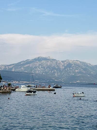 This media file shows a scenic view of a body of water with several boats docked and people enjoying leisure activities. In the foreground, a concrete pier lines the water's edge, with numerous individuals gathered, some seated and others standing. Various boats, including a sailboat and several motorboats, are moored to the pier. One motorboat is white with a dark hull, and another appears to be a smaller, open vessel. Further out in the water, a white boat with a dark roof is visible, with a person swimming nearby.
Dominating the background is a rugged, mountainous landscape. The slopes are covered in vegetation and show signs of rocky terrain. A town or village is nestled at the base of the mountains, with buildings clustered along the coastline.
The time of day appears to be daytime, possibly late afternoon, given the soft lighting and the angle of the sun. The weather seems pleasant, with a clear blue sky and some wispy white clouds. The water is calm with gentle ripples, reflecting the light. The overall atmosphere suggests a relaxed holiday or vacation setting. There is no visible text in the media file. The location context provided is Đenovići, Montenegro.