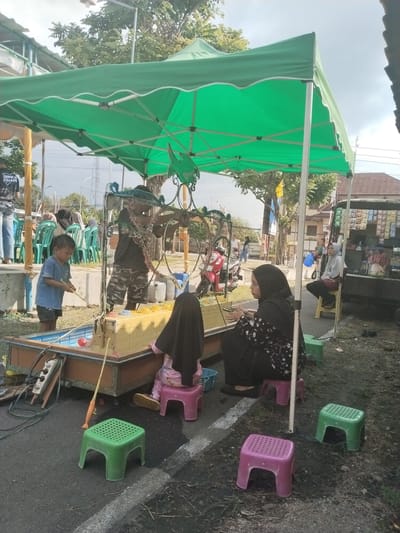 In Sambirejo, Indonesia, a daytime street scene shows a fair game of "fishing" set up under a green canopy. A small, rectangular pool filled with water and colorful floating objects sits in the center. A young boy in a blue shirt and shorts stands by the pool, holding a fishing rod, presumably trying to "catch" the objects. A woman in a black hijab sits on a pink stool beside the pool, watching a young child in pajamas also sitting on a pink stool. A man in a black shirt and camouflage shorts stands behind the pool, possibly overseeing the game. Several green and pink plastic stools are scattered around the area. To the right, a snack food vendor sits near their vehicle, and in the background, there is a row of green plastic chairs and other passers-by. A motorcycle and a building are also visible in the background. The game is set up on the street.