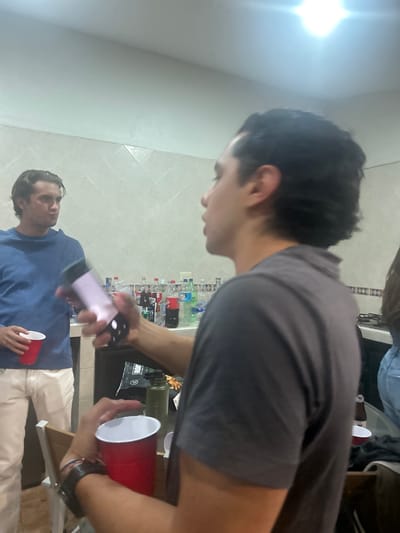 A group of people are indoors, likely at a social gathering. One man is in the foreground, wearing a grey shirt and holding a red plastic cup. Another man is holding a phone with its camera activated in the direction of the man with the cup. He is wearing a blue shirt and white pants, and also holding a red cup. The background has a tiled wall with a countertop covered with water bottles and other objects. It is unclear where in San Juan Cuautlancingo, Mexico this image was taken.