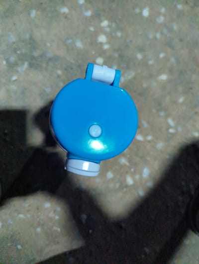 A top-down, medium shot shows the lid of a blue water bottle with white accents, resting on a textured, light brown surface that appears to be concrete or asphalt. The bottle lid is round and smooth, with a slightly raised, lighter blue circular button in the center. There are white, cylindrical pieces attached to the sides of the lid, suggesting a cap mechanism.
The background is out of focus, but it shows a mottled texture with small, lighter specks scattered across a darker, shadowed area. The lighting suggests it might be daytime, with a shadow falling across the right side of the scene, possibly from an unseen object or structure. The overall impression is of an everyday object placed outdoors. No people are visible, and no text is discernable on the bottle or in the surrounding environment. The location context provided is Elele Alimini, Nigeria.