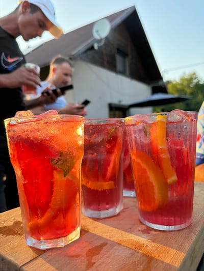 In Mordarka, Poland, four glasses of red-hued beverages are arranged on a wooden surface in the foreground. The glasses are filled with ice, slices of orange, and what appears to be mint leaves, suggesting a refreshing drink. The sunlight, casting warm golden light, glints off the condensation on the glasses, indicating a bright, likely warm, outdoor setting. In the background, two men are visible. The man on the left, wearing a white cap and a black t-shirt with a Nike logo, holds a glass of a similar red beverage. The man to his right, in a blue and white striped shirt, is looking at his phone. Behind them, a building with a wooden facade and a brown roof, possibly a house, is partially visible, with trees and foliage indicating a suburban or rural environment. The overall scene depicts a casual gathering outdoors on a sunny day.