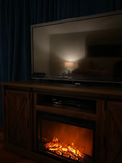 The image displays an interior scene in Ridgefield, United States, centered on a home entertainment setup. A dark-screen Panasonic flat-panel television rests on a dark-stained wooden media console. The console features two side cabinets with X-patterned, sliding barn-door style fronts. A central open compartment within the console houses a black Samsung Blu-ray player. Below the Blu-ray player, an operational electric fireplace insert exhibits artificial glowing logs and simulated orange flames. To the left of the television, dark blue, pleated curtains hang vertically. The television screen reflects an illuminated table lamp with a light-colored shade, a wall, and blurred dark shapes resembling furniture, indicating the room's opposite side. No individuals are present. The primary light sources are the electric fireplace and the reflected lamp, casting a dim illumination over the immediate area.
