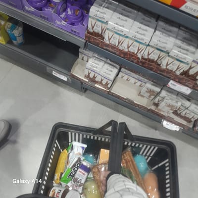 This image appears to have been taken inside a grocery store in the city of Nuseirat, Palestinian Territory. It shows a shelf stocked with chocolate and candy products, including brands such as Milka and Amada. A black shopping basket with various items is also visible, including snacks like Todo and Kremali, and other packaged goods. The scene depicts a shopper presumably selecting items from the shelf. The "Galaxy A14" watermark suggests the image was taken using that specific model of phone.