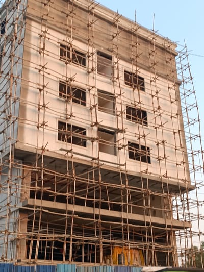 A multi-story residential or commercial building in Anantapur, India, is depicted under active construction. The structure, observed from a low-angle perspective, features at least five visible floors, with the upper sections exhibiting a white, plastered facade and dark-framed rectangular window openings. The topmost floor includes a decorative cornice. The entire visible exterior of the building is enveloped in an extensive, multi-level scaffolding system constructed from numerous interconnected bamboo poles secured horizontally and vertically. The lower floors and ground level are partially obscured; the immediate foreground reveals a barrier composed of blue and light blue corrugated metal panels, behind which yellow tarpaulins are visible, possibly covering materials or a temporary shelter. No individuals, specific actions, or interactions are visible. The background consists of a clear, light blue sky, indicating daylight conditions.