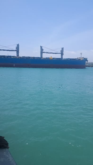 The image displays a large cargo ship, consistent with a bulk carrier, floating on calm, teal-blue water under a clear, light blue sky. The vessel's hull is dark blue, with a distinct maroon section below the waterline. At least four grey-colored deck cranes, equipped with visible rigging, are positioned along the ship's deck, with minor yellow structural components also discernible. The ship appears to be either stationary or moving slowly within a port or channel. A dark, possibly metallic, edge of an unknown structure or another vessel is partially visible in the bottom-left foreground. No individuals, specific actions, or direct interactions are visible. This maritime scene is consistent with a port environment in the Ganjoni/Shimanzi ward of Mombasa, Kenya.