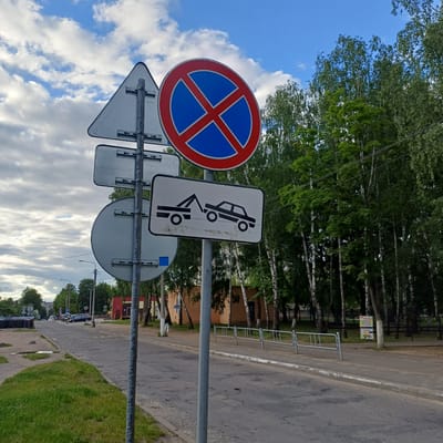 The image captures an urban street scene in Baranavichy, Belarus, centered around a cluster of road signs. In the immediate foreground, two vertical metal poles are positioned on the edge of a cracked asphalt road and a patch of unkempt green grass with a small puddle.
The leftmost pole supports the rear sides of three white, non-reflective signs: a triangular sign at the top, a rectangular sign below it, and a circular sign at the bottom. The rightmost pole displays two prominent signs facing the viewer. The upper sign is a circular "No Stopping" (or "No Halting") sign (Belarusian road sign 3.27), characterized by a blue disk with a thick red border and a diagonal red 'X' intersecting its center. Directly beneath it is a rectangular white supplementary sign with a black border, featuring a black silhouette of a tow truck actively towing a passenger car, indicating a "Tow Away Zone."
The asphalt road, showing numerous cracks and patches, extends into the midground. Yellow transverse markings, possibly speed bumps or road humps, are visible across the road further down. On the right side of the road, a concrete curb separates the main thoroughfare from a paved sidewalk, which is lined with a series of silver-colored metal pedestrian barriers or bicycle racks.
The right side of the frame is dominated by a dense line of deciduous trees with vibrant green foliage, many displaying characteristic white birch bark, creating a natural screen against the distant sky. Faint utility lines are visible traversing the upper right section among the treetops. A colorful poster is situated on a pole or structure behind the metal barriers in the far right background, adjacent to a dark fence.
In the background on the left, several parked or stationary vehicles are visible along the roadside, including a dark sedan. Beyond these, a mix of architectural structures can be observed: distant multi-story residential buildings (apartments), a light-colored, possibly brick, building, and a smaller, red-painted building with a dark roof closer to the midground right.
The sky is partly cloudy, featuring patches of clear blue interspersed with white and grey cumulus clouds. No people or active interactions are discernible within the frame, contributing to a sense of a quiet, functional urban environment.