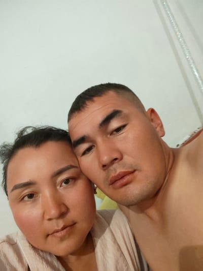 This is a close-up, selfie-style photograph of two individuals, a man and a woman, taken indoors in Ylay-Talaa, Kyrgyzstan. The man is on the right, with his head tilted slightly, and the woman is on the left, her head resting close to his. Both appear to be of Asian descent. The man has a shaved head and dark eyebrows. The woman has her hair tied back loosely and prominent eyebrows. They are both looking towards the camera with neutral expressions. The background consists of a plain white wall, with a hint of a decorative, possibly crystal, element visible on the upper right side of the frame. The lighting is soft and appears to be from an artificial source. There is no indication of the exact location within Ylay-Talaa, such as a specific street or landmark. The image focuses on the faces and upper torsos of the two subjects.