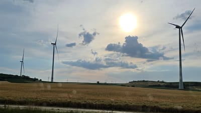 The image depicts a landscape in Bullendorf, Austria, featuring a field of golden wheat in the foreground, with rolling hills and numerous wind turbines in the background. The sun is prominently visible in the upper right portion of the sky, partially obscured by clouds. Several wind turbines are scattered across the landscape, with two larger turbines positioned on the right and left sides of the image, and smaller ones visible in the distance. The sky is a mix of blue and grey clouds, with some lens flares visible. The terrain appears to be agricultural, with vineyards or crops visible on some of the hillsides. The overall scene conveys a sense of rural, open space with a focus on renewable energy infrastructure.