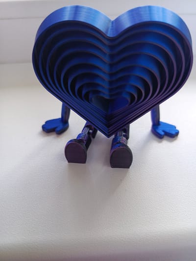 This is a close-up, eye-level shot of a blue, heart-shaped object with legs and feet, standing on a white, textured surface. The object appears to be 3D printed, with visible layering lines, especially on the legs and feet. The heart shape is composed of multiple concentric heart outlines, creating a layered effect. The color is a deep, iridescent blue that shifts to purple in some areas due to the lighting. The object has two simple, stylized blue feet and two articulated legs, one of which is partially obscured. The background is a bright, plain white, suggesting an indoor setting. The lighting is even and bright, casting subtle shadows. There is no text visible. The overall mood is whimsical and playful, likely representing a decorative item or a toy. The context of Biaroza, Belarus, suggests a potential origin or ownership location for this object.