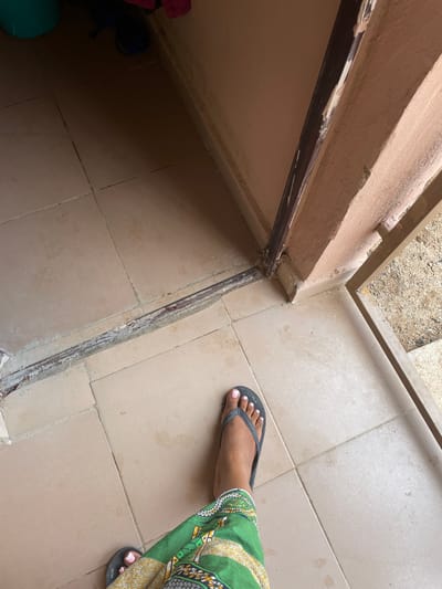 The image depicts a floor area with light brown square tiles. A dark brown door frame is visible, with some paint chipping on its surface. A person is wearing a green and yellow patterned skirt and grey flip-flops, with painted toenails. The scene appears to be indoors, possibly a doorway or an entrance area. The floor is somewhat dusty, and there are some water marks visible. The overall impression is of an unkempt, possibly abandoned, or lived-in domestic space. The location is specified as Zanzi, Nigeria.