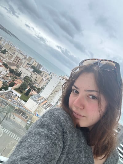 A young female with brown hair, wearing a grey knitted sweater, is depicted in the foreground, looking directly at the camera. Tortoiseshell sunglasses are positioned on top of her head. The scene is captured from a high vantage point, overlooking the city of Les Meravelles, Spain. The urban environment below features numerous multi-story buildings, predominantly light-colored, with some exhibiting red-tiled roofs, and interspersed palm trees. A street with a zebra crossing and a white vehicle is visible directly below. Beyond the city, the sea extends to the horizon, with waves visible near the shoreline and a distant landmass on the far left. The sky is entirely overcast with dense grey clouds.