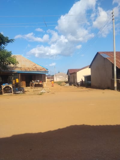 This is a daylight, eye-level shot of a dusty street in Mangu Town, Nigeria, with a few buildings lining the road. The sky is bright blue with scattered white clouds. On the left, a long, single-story building with a rusty corrugated iron roof has a blue trim along its overhang. In front of this building, there are wooden tables with various items, including yellow containers, and a blue motorcycle. Further down the street, to the right, are two more buildings. The first is a lighter-colored structure with a similar rusty corrugated iron roof, and the second is a larger, beige building with a dark entrance. A person in a yellow shirt and orange pants is walking away from the camera on the right side of the street. Another person in a dark shirt and pants stands near a motorcycle in front of the second building on the right. Power lines stretch across the upper portion of the image, supported by a utility pole on the far right. The ground is a sandy, reddish-brown dirt. A shadow of an unseen object falls across the foreground.