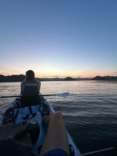The image captures a serene sunset scene on Skadar Lake, near Vranjina, Montenegro. Two people are in a kayak on the calm water. The person in the front, facing away from the camera, is wearing a life vest and appears to be looking towards the horizon. The back of the kayak shows the legs and feet of another person, wearing Crocs, suggesting they are the photographer. The sky is painted with soft hues of blue, orange, and pink, transitioning into dusk. Distant, dark mountains form a silhouette against the colorful sky. The water reflects the sky's colors, creating a tranquil and picturesque atmosphere.
