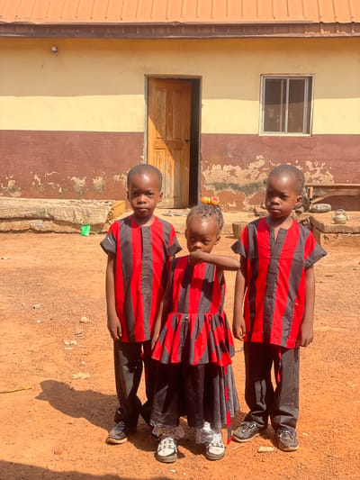 Three young children are depicted standing outdoors under bright sunlight in Oturkpo, Nigeria. The central figure is a girl, flanked by two boys, all appearing to be of African descent. All three wear coordinating red and black striped attire; the boys in short-sleeved tunics and dark trousers, the girl in a dress over dark trousers and white lace-trimmed leggings. The girl's braided hair is adorned with colorful beads, and her right hand is raised towards her mouth. They stand on unpaved reddish-brown earth, which shows distinct shadows. The background consists of a single-story building with a reddish-brown corrugated metal roof, a cream-colored upper wall section, and a deteriorating dark brown lower wall section. A wooden door, slightly ajar and casting a shadow, is centrally located, with a metal-framed window to its right. A small light fixture is present on the wall above the door. A low, unfinished concrete or earthen ledge runs along the building's base. Various household items, including a silver bowl, a dark pot, and a light-colored kettle, are visible on the ground to the right of the building. A light green plastic bottle or wrapper is visible on the ground to the left.