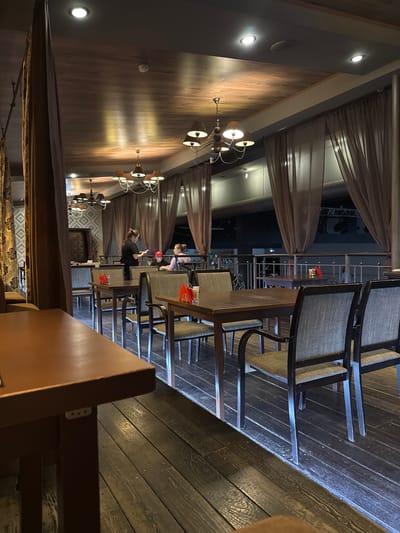 The image depicts the interior of a restaurant in Balezino, Russia. The dining area is lit by chandeliers and recessed ceiling lights, creating a warm ambiance. The walls are paneled with wood. The room is furnished with wooden tables and chairs with woven seats. Some tables are occupied by patrons. There are brown drapes along the side of the room. A metal railing is visible outside, beyond the drapes.