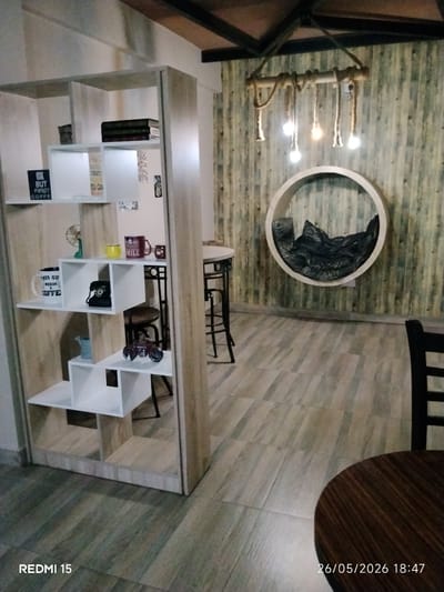 This is an interior shot. A shelving unit is filled with mugs, books, and decorative items like a small fan and a toy car. There is a table with bar stools partially visible behind the shelf. In the background, a wall with a wooden texture is adorned with a circular structure filled with a dark material, possibly cushions or fabric. Above the wall decoration, a rope-like light fixture with exposed bulbs hangs. A wooden table with a chair is in the right foreground. The floor has a light wood grain pattern. The text "REDMI 15" and "26/05/2026 18:47" are present at the bottom of the image, suggesting it was taken on a Redmi 15 device with the date and time stamp. There is no indication of location in the city of Kabala, Nigeria.