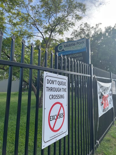 The image captures a fence lining the perimeter of a school, likely Middle Park State School, located in Riverhills, Australia. The scene is outdoors during daylight hours, indicated by the bright sky and natural lighting, with clear weather and some scattered clouds. Lush green grass is visible on the school side of the fence, along with mature trees that cast some shade.

Prominently displayed on the black metal fence are several signs and a banner. A blue and white sign, featuring the school's logo, identifies it as "Middle Park STATE SCHOOL" with the motto "Each to Succeed." Below this, an electronic display shows the message "Happy Holidays," suggesting a school break or upcoming holiday period.

To the right of the school sign, a white rectangular sign with black and red text delivers a clear safety message: "DON'T QUEUE THROUGH THE CROSSING." Below this, a large red circle with a diagonal line through it signifies "NO EXCUSES," with the smaller text "A message from your kids" positioned beneath. This sign aims to encourage safer traffic practices around the school crossing.

Further along the fence, a white banner is tied, featuring a red graphic of an open hand in a "stop" gesture and the word "Stay" partially visible in black text, likely conveying a broader safety message related to staying safe. There are no people visible in the image, making the focus entirely on the school's entrance area and its informational and safety signage.