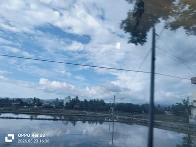 The image shows a view from inside a vehicle, likely a bus or car, through a window. The scene outside is a rural landscape in Bandasafa, Indonesia, as indicated by the camera timestamp. In the foreground, there are rice paddies flooded with water, reflecting the sky. Beyond the rice paddies, there is a line of trees and vegetation, with some houses interspersed. In the background, there are distant hills or mountains. Overhead powerlines run across the sky. Reflections are visible on the window: a blurred view of trees, and also of the interior. A utility pole partially obscures the view on the right side of the frame. The sky has scattered clouds and blue patches.