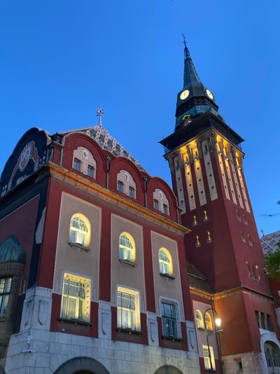 This media file captures a striking view of the Subotica City Hall in Serbia, an iconic Art Nouveau architectural masterpiece, during the twilight hours. The image is taken from a low, upward angle, showcasing the building's intricate details against a clear, deep blue sky.
The main building features a multi-tiered facade, primarily in a rich reddish-brown, contrasted with sections of light, possibly pale pink or beige, plasterwork surrounding its numerous windows. The architecture is highly decorative, with elaborate white and grey stucco or carved patterns adorning the window frames and the complex, ornamental roofline, which includes elements with a mosaic-like texture. Many of the windows, both arched and rectangular with multiple panes, are brightly illuminated from within, casting a warm glow that stands out against the cool evening sky.
A tall, slender clock tower dominates the upper left of the image. Its lower section shares the red-brown color and horizontal lighter stripes of the main building, transitioning to a darker, possibly oxidized metal, upper part that houses several illuminated clock faces. The spire of the tower is pointed and topped with a cross or finial. One clearly visible clock face indicates a time close to 8:40. Artificial streetlights are also visible, contributing to the evening ambiance.
The bottom right corner reveals a section of geometrically patterned pavement. No people or specific events are depicted; the focus is entirely on the building's grandeur and its distinctive, ornate architectural style. The scene conveys a sense of historical beauty and intricate craftsmanship. No discernible text is visible on the building's exterior.